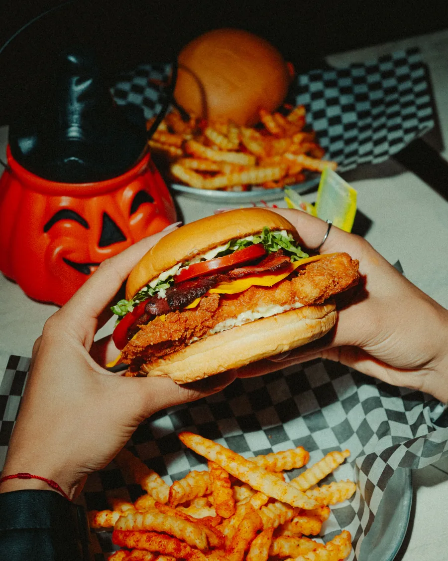Dos manos sosteniendo una hamburguesa de pollo crocante sobre una bandeja de papas