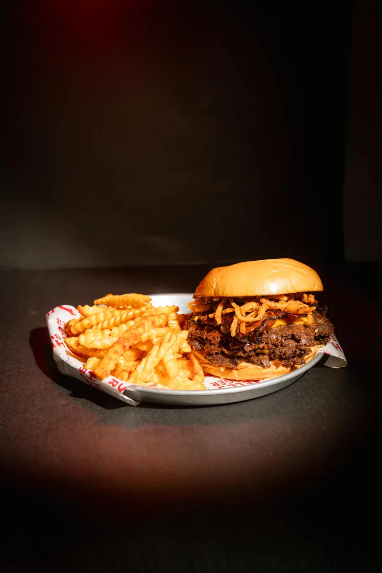 Una hamburguesa con cebolla crocante y papas fritas onduladas, servidas en un plato con papel de la marca