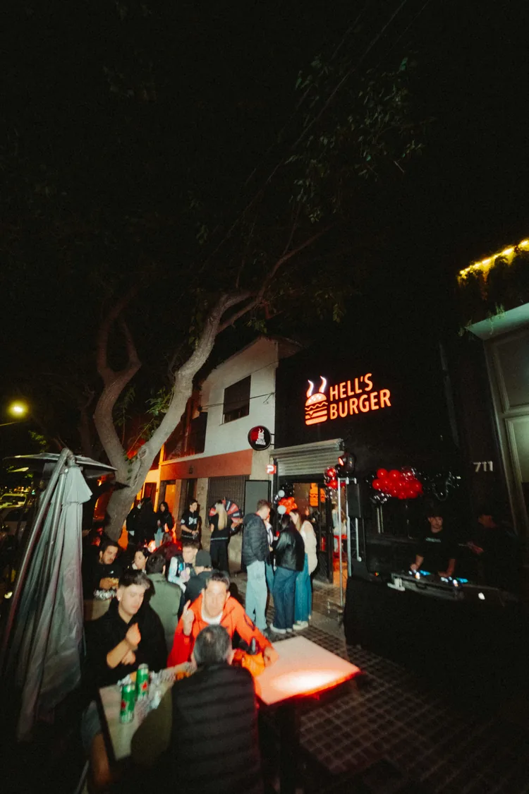 La fachada del local de noche, con el cartel de Hell’s Burger encendido y gente esperando en la vereda
